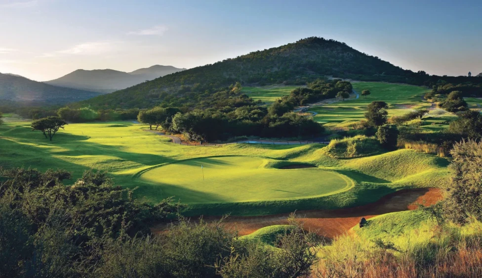 Green del Lost City Golf Club rodeado de colinas, vegetación y luz cálida.
