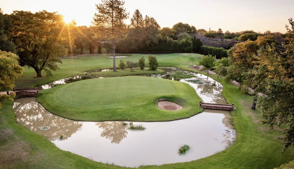Green del Pretoria Country Club rodeado de agua, un búnker y árboles al atardecer.