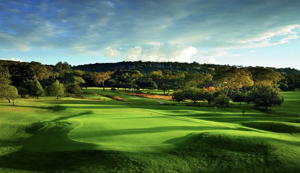Green del Royal Johannesburg Country Club con colinas, árboles y cielo dramático al fondo.